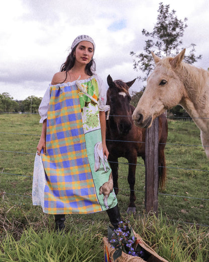 A woman stands in a grassy field with two horses behind a wire fence, wearing a colorful patchwork apron dress with blue, yellow, green, and white plaid sections and animal illustrations. A basket of flowers sits on the ground in the foreground.