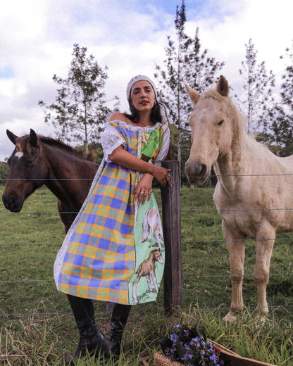 A woman wearing a colorful plaid and printed dress stands outdoors beside a fence, with a brown horse on her left and a white horse on her right. The dress features a yellow and blue plaid pattern with a green panel showing goat illustrations.