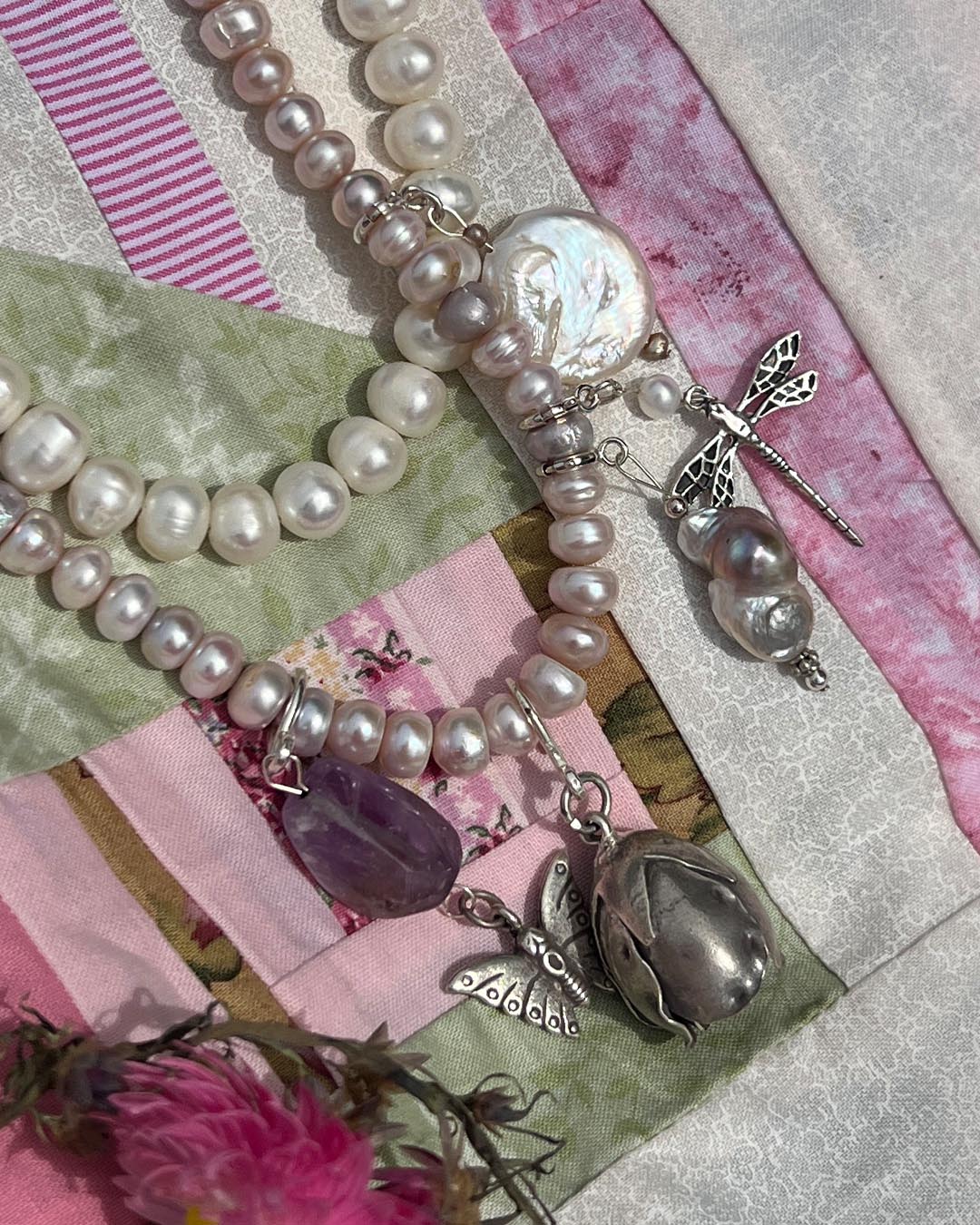 A close-up of a layered pearl necklace with mixed white and pale pink pearls, silver-toned metal charms including dragonflies and a flower-shaped pendant, and a purple amethyst bead, styled on a patchwork fabric background with pink ribbons and dried flowers.
