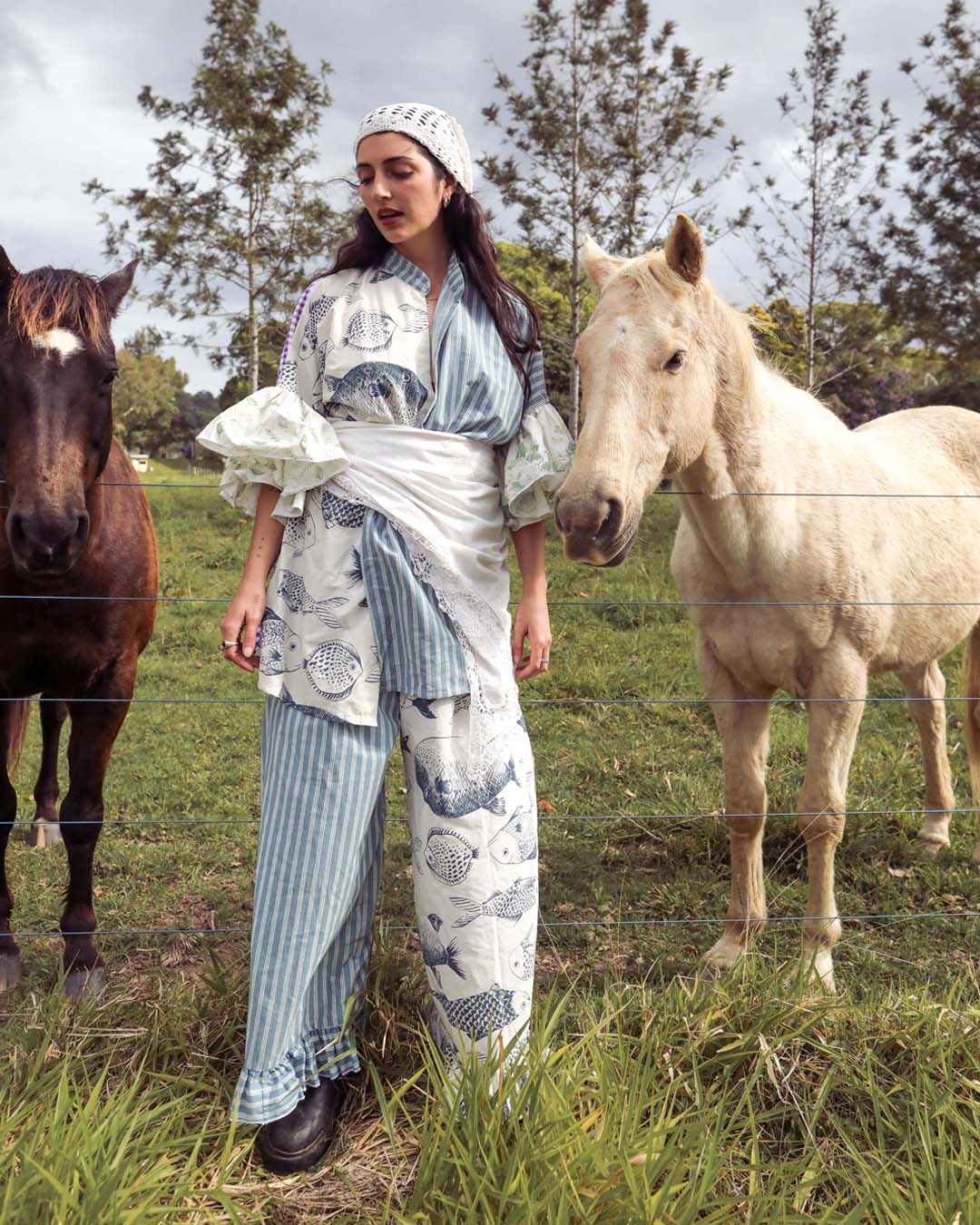 A woman stands outdoors in a grassy field between two horses, wearing a matching blue-and-white patterned outfit with a white headscarf. The clothing features vertical stripes and fish motifs, and the scene is photographed in natural daylight.