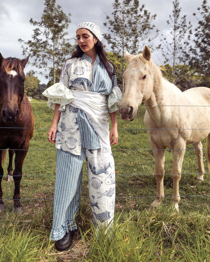 A woman stands outdoors in a grassy field between two horses, wearing a matching blue-and-white patterned outfit with a white headscarf. The clothing features vertical stripes and fish motifs, and the scene is photographed in natural daylight.