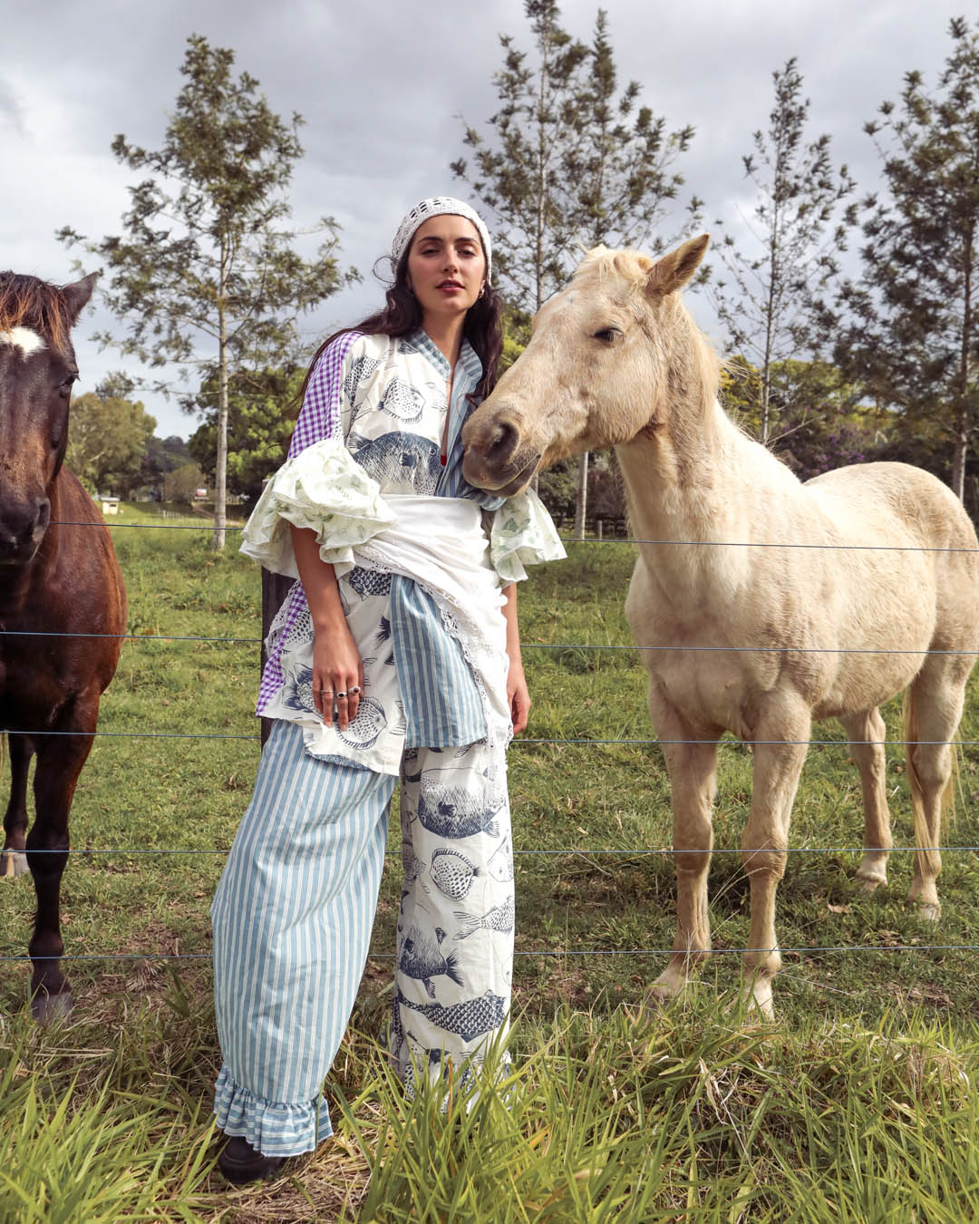 A woman stands outdoors in a grassy field between two horses, wearing a layered outfit with a purple patterned shirt, white ruffled sleeves, a white apron, and wide-leg striped pants with blue fish illustrations.