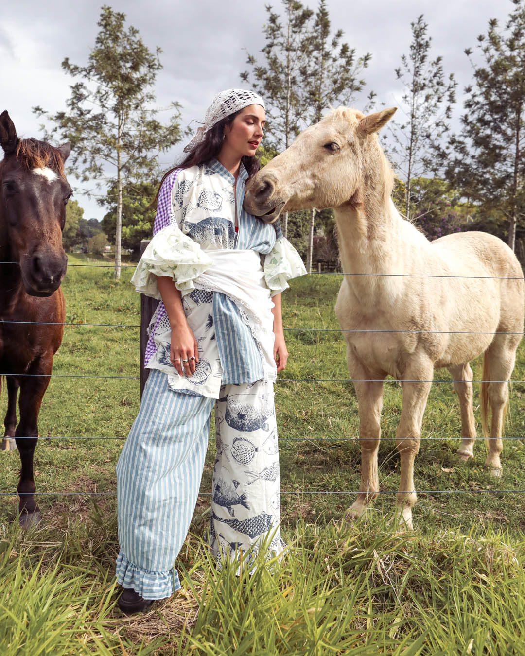 A woman wearing a bohemian-style printed outfit stands in a grassy field between two horses, with trees and an overcast sky in the background.