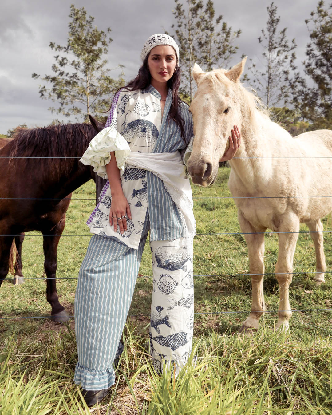 A woman wearing a matching blue-and-white patterned outfit stands outdoors in a grassy field between two horses, with one horse touching her hand. The outfit includes a white top with blue floral motifs, blue-and-white striped pants, and a white head covering.