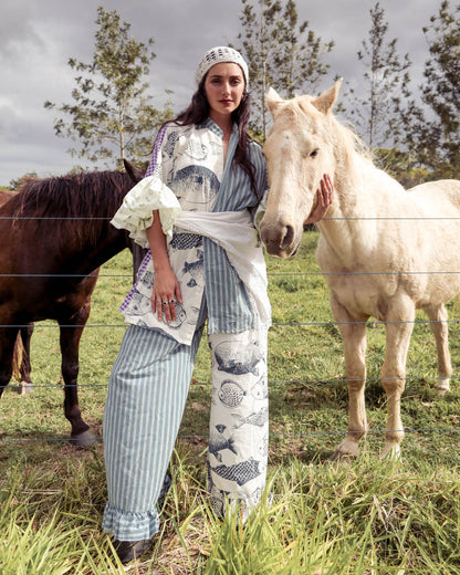 A woman stands between a dark coloured horse and a light coloured horse. She is wearing one-of-a-kind patchwork bohemian handmade shirt smock dress that is tied at the waist with an heirloom lace apron. She also wears matching mix match patchwork pants.