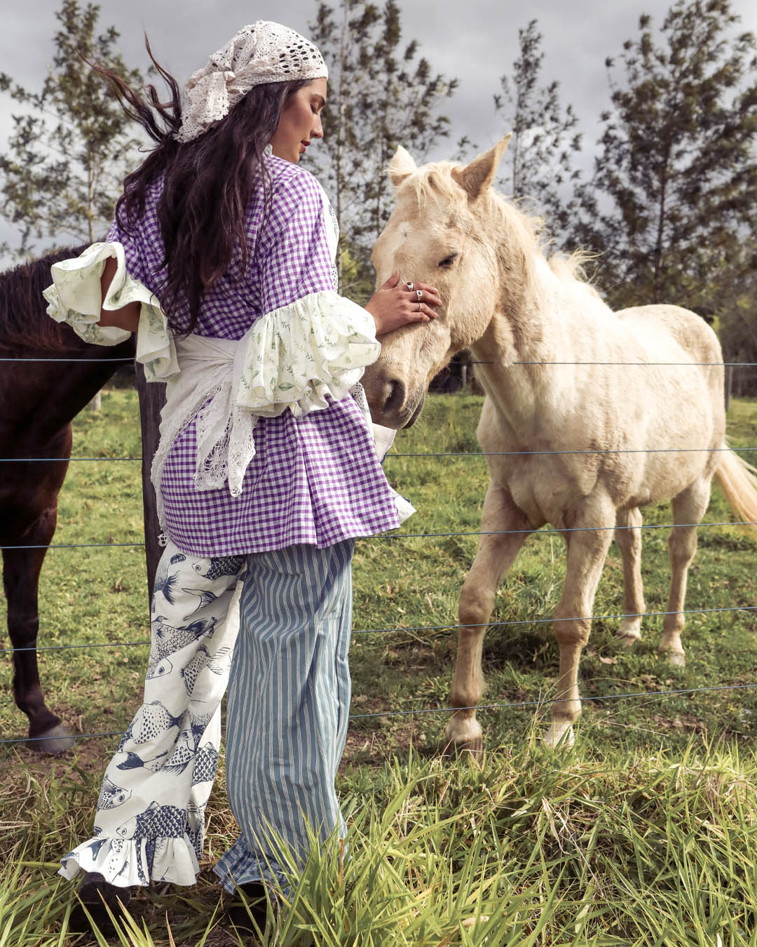 A woman in a purple gingham blouse, white apron, and striped pants stands in a grassy field, touching the head of a light-colored horse while another dark horse is visible behind her.