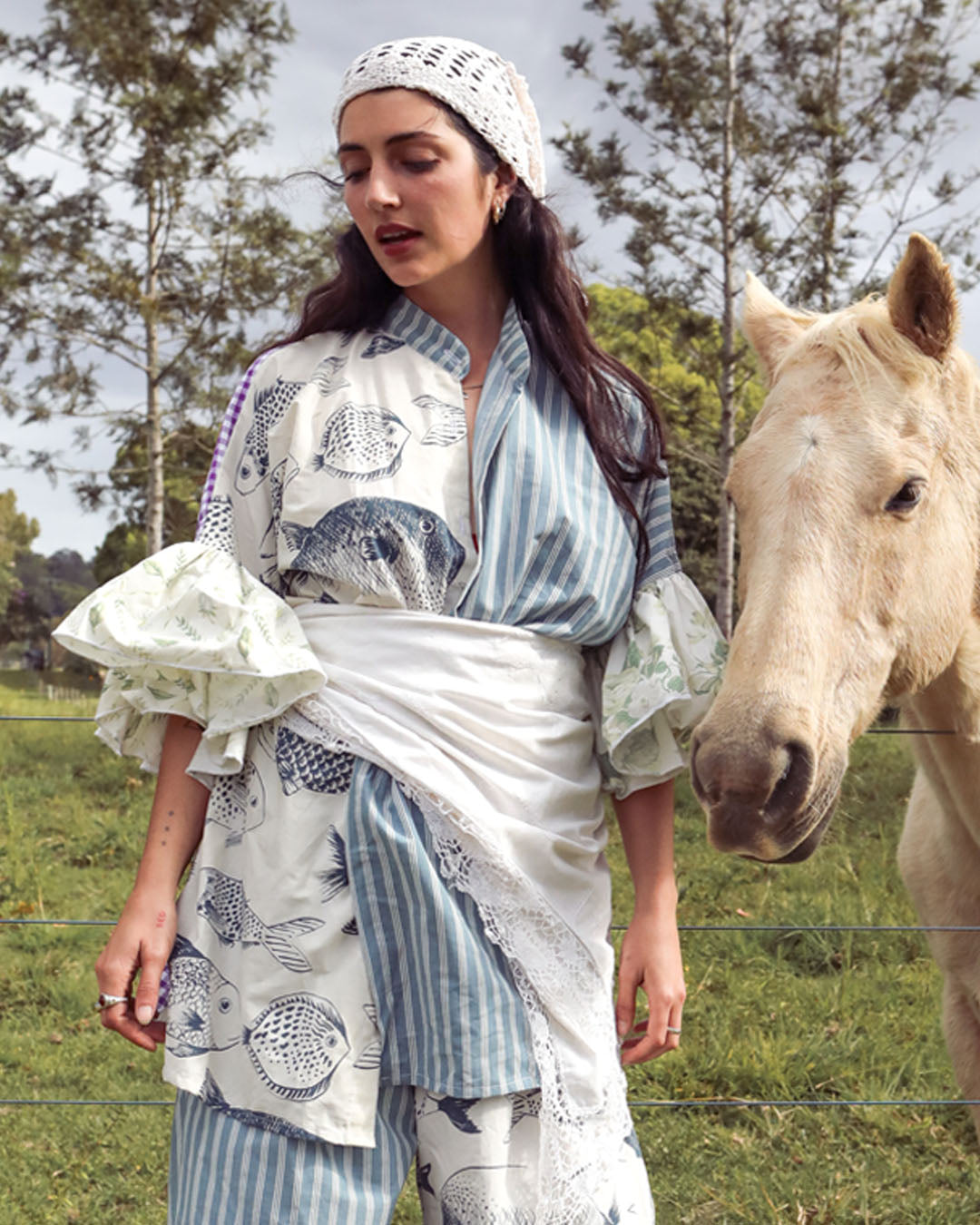 A woman wearing a blue and white striped kimono-style top with fish prints, a matching headscarf, and striped trousers stands outdoors next to a light-colored horse in a grassy field.