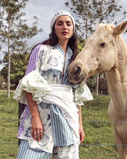 A woman wearing a bohemian-style patchwork outfit with blue and white stripes, white lace, and purple gingham stands outdoors next to a light-colored horse in a grassy field.