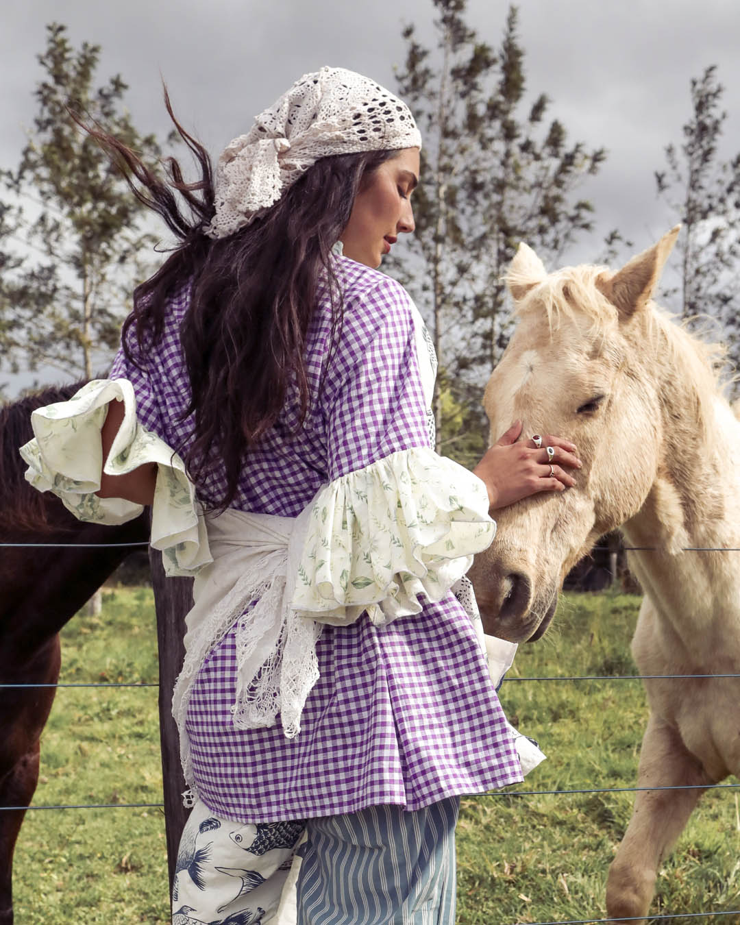 A woman in a purple and white gingham dress with lace sleeves stands in a grassy field next to a light-colored horse, touching its face. She wears a white lace headscarf.