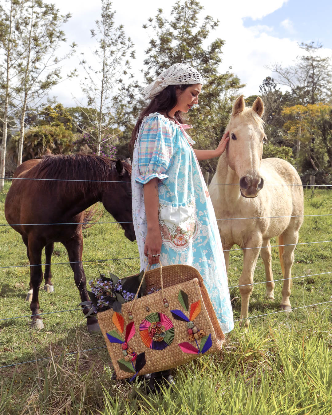 A woman in a light blue and white patterned dress stands in a grassy field with two horses, holding a large woven straw tote bag decorated with colorful floral patches.