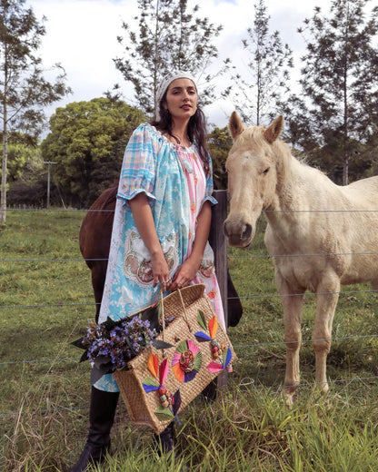 A woman wearing a light blue and pink patterned dress stands in a grassy field next to a white horse, holding a woven tote bag decorated with colorful floral embroidery and purple flowers.