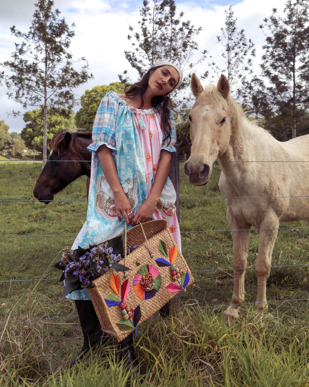 A woman stands in a grassy field with two horses behind a wire fence, wearing a light blue and pink patterned dress and holding a large woven tote bag decorated with colorful floral and leaf motifs.