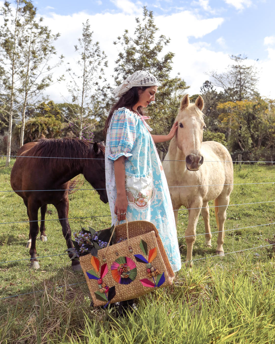 A woman wearing a light blue and white patterned dress and a white lace headpiece stands in a grassy field next to a light-colored horse, holding a woven straw basket decorated with colorful butterfly and floral patterns. Another dark horse is visible in the background.