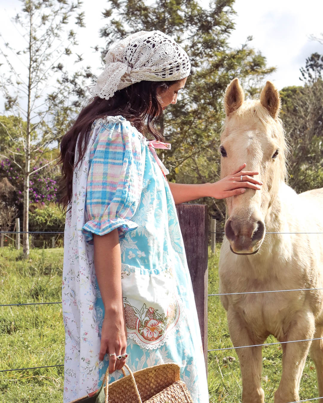 A woman in a light blue and white floral dress with a lace headscarf pets a light-colored horse in an outdoor field.