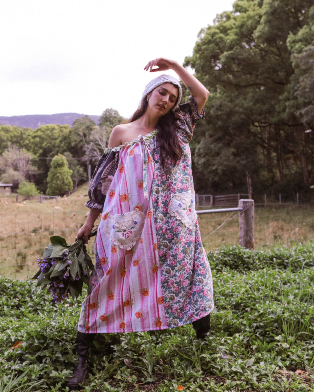 A woman stands in a grassy outdoor field wearing a one size, one of a kind, loose, off-the-shoulder patchwork dress with floral and striped panels, a white headscarf, and dark boots, while holding a bouquet of green leaves.