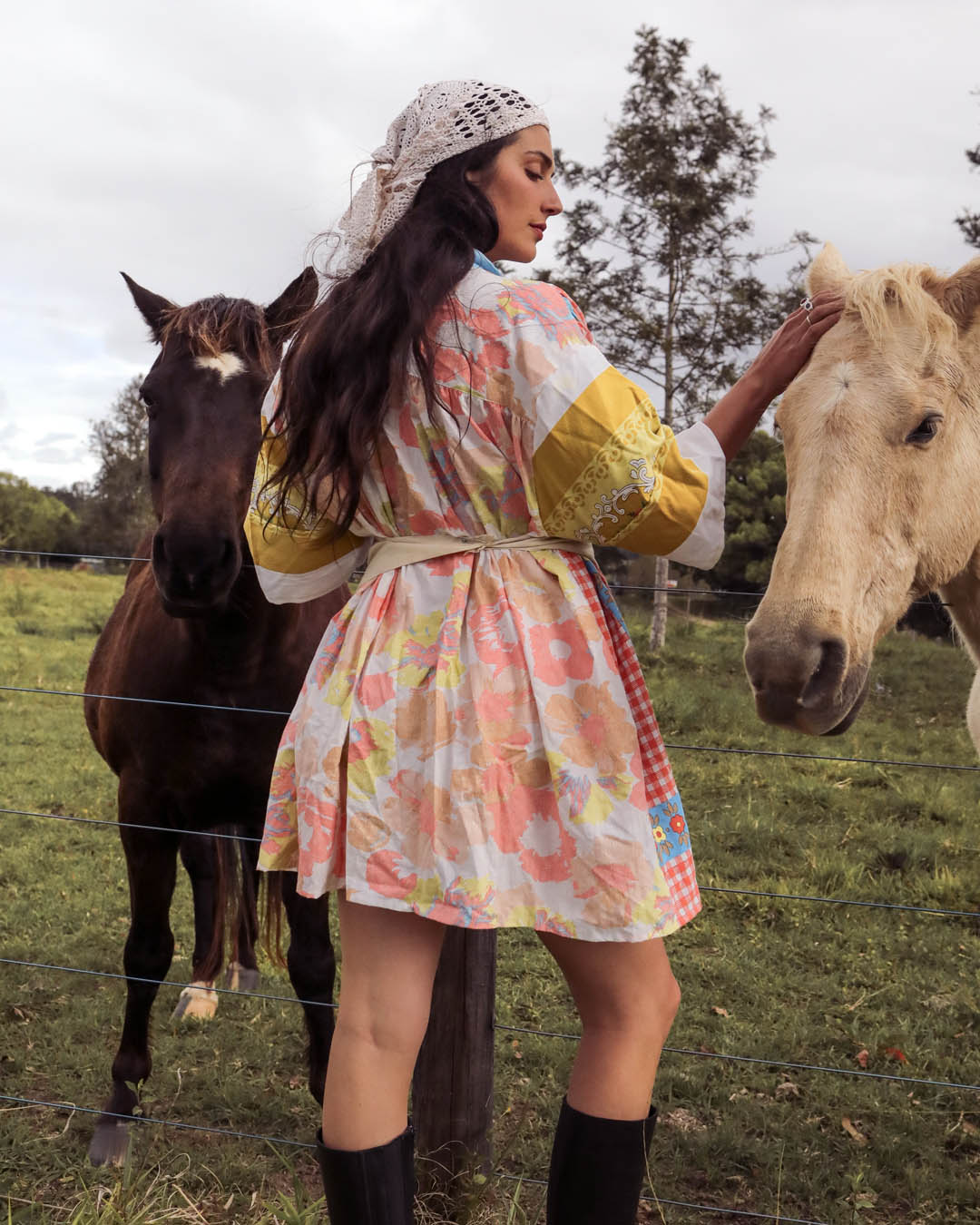 A woman wearing a colorful floral print kimono-style dress stands in a grassy field, touching a light-colored horse while a darker horse stands behind her. The dress has yellow sleeves and a multicolor pastel floral pattern.