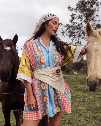 A woman wearing a colorful patchwork kimono-style robe stands outdoors in a grassy field with two horses nearby. The robe features a light blue front panel, red-and-white gingham sleeves and hem, yellow shoulder panels, and a beige sash with floral embroidery.