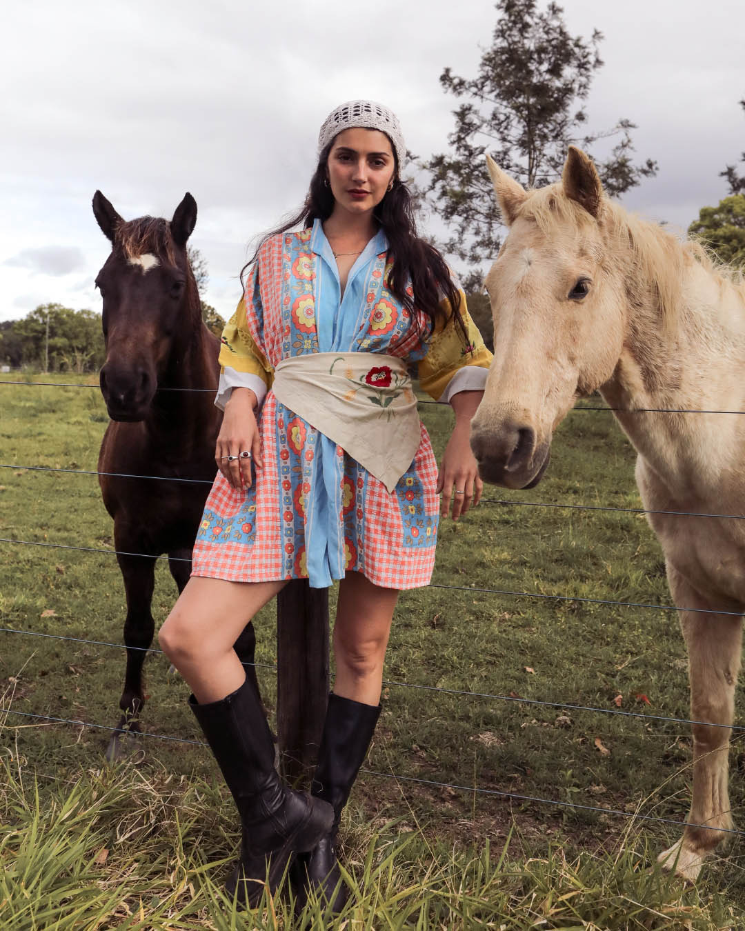A woman stands outdoors in a grassy field between two horses, wearing a colorful patterned dress with a light blue front panel, a beige sash, a white headband, and black knee-high boots.