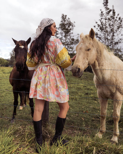 A woman wearing a colorful floral dress, black boots, and a white headscarf stands in a grassy field between two horses, reaching out to pet the light-colored horse on the right.