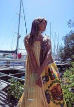 Load image into Gallery viewer, Woman in a colorful dress standing by a marina with boats and trees in the background