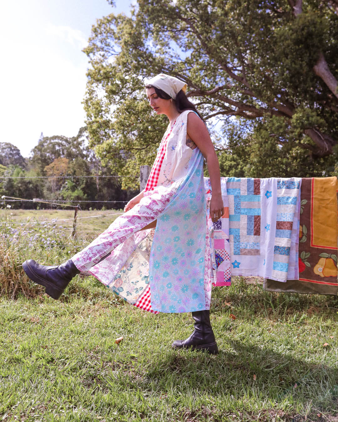 A woman wearing a long sleeveless dress with a floral print and a red-and-white gingham underlayer walks outdoors in a grassy field with trees and a quilt hanging in the background.