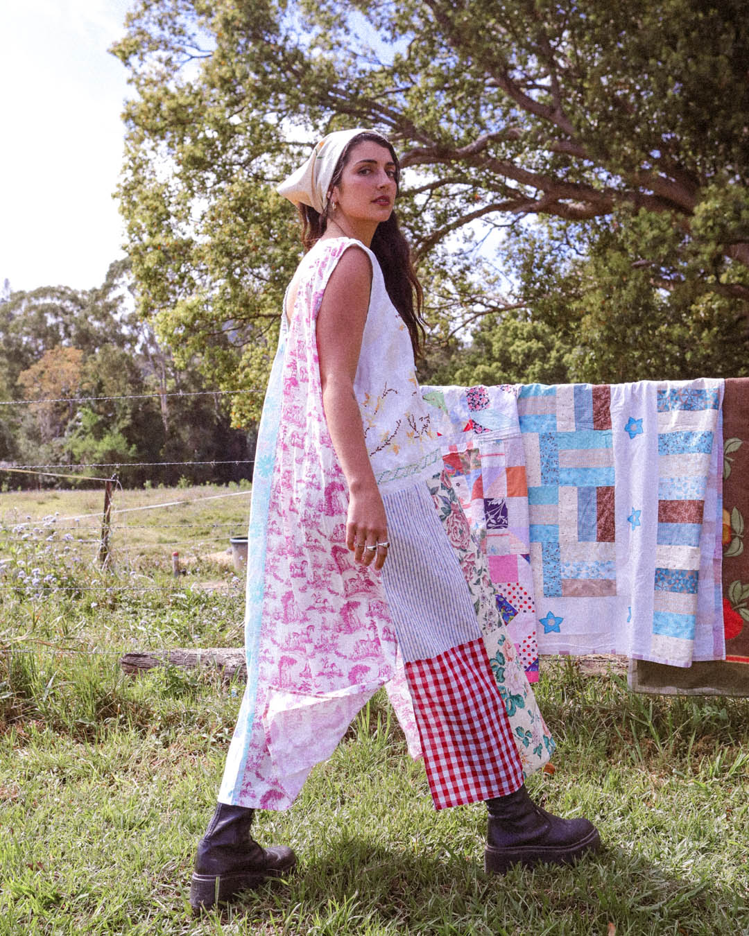 A woman stands outdoors in a grassy field wearing a long, patchwork-style dress with a white base and pink floral print, red gingham sections, and a white apron-style overlay. A colorful quilt hangs on a line behind her, with trees in the background.