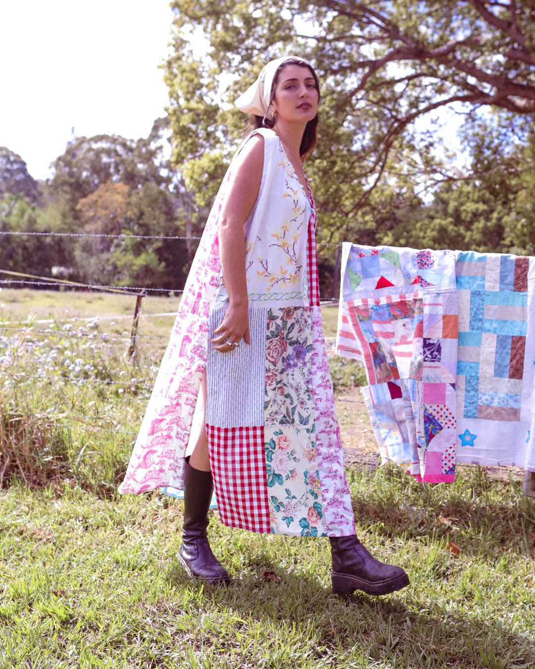 A woman stands outdoors in a grassy field wearing a long patchwork dress with floral, gingham, and knit panels. A matching patchwork quilt hangs on a clothesline in the background, with trees and a fence behind her.