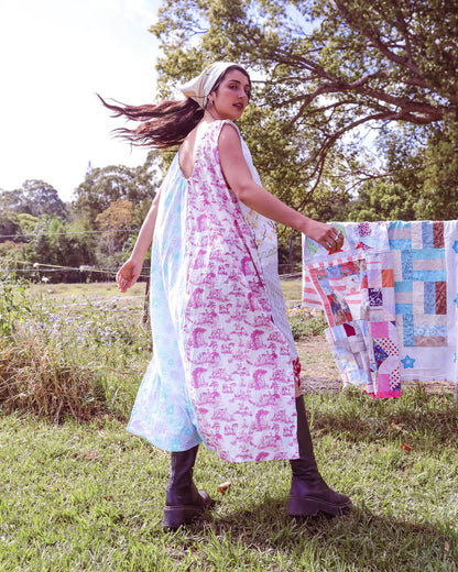 A woman outdoors in a grassy yard wearing a long sleeveless dress with a split design: one side is light blue and the other is white with pink floral print. She is holding up a colorful patchwork quilt next to her.