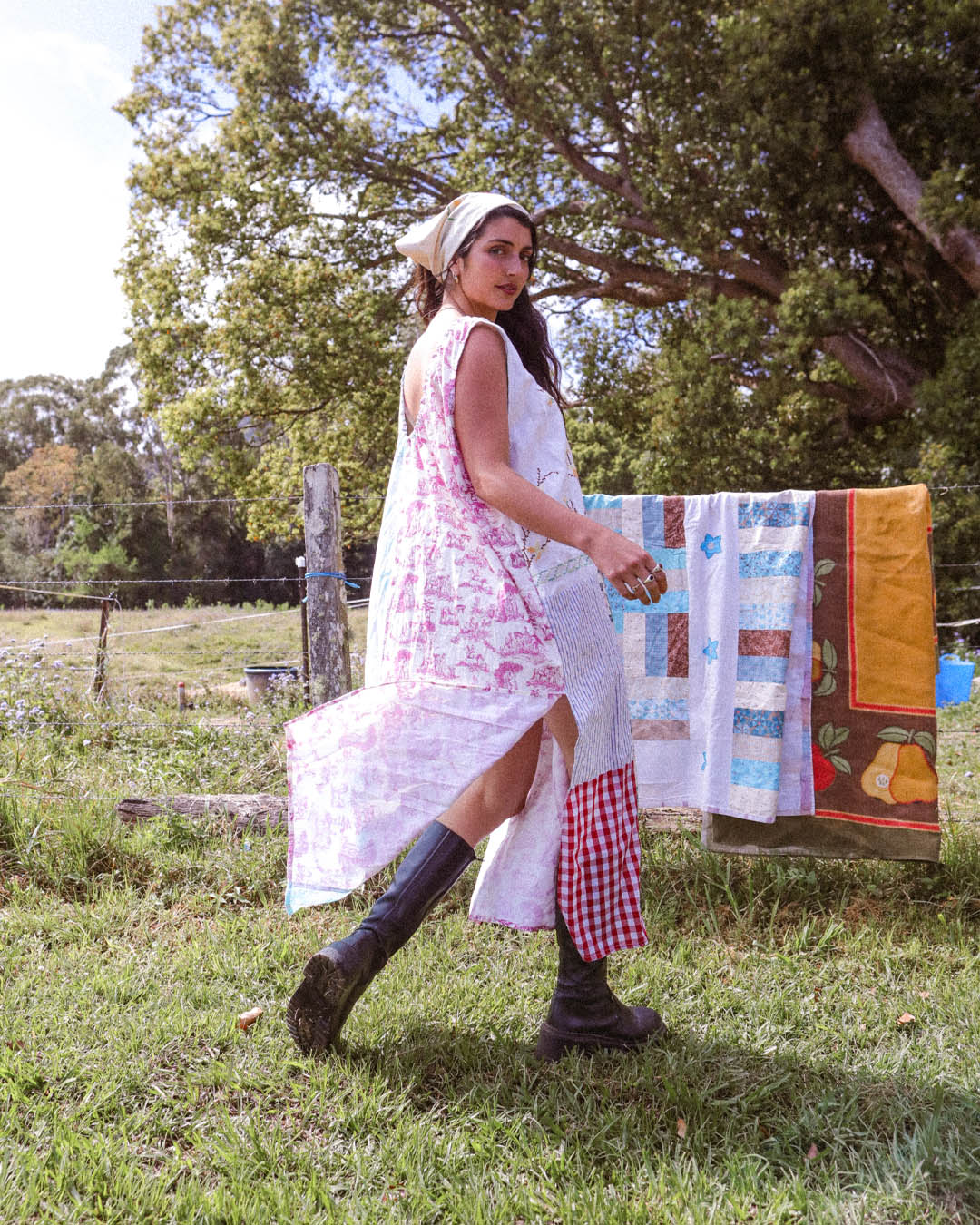A woman wearing a long white dress with pink floral print and a matching headscarf walks outdoors in a grassy field. A colorful patchwork quilt is hanging on a clothesline in the background under large trees.