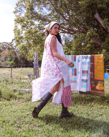 A woman wearing a long white dress with pink floral print and a matching headscarf walks outdoors in a grassy field. A colorful patchwork quilt is hanging on a clothesline in the background under large trees.