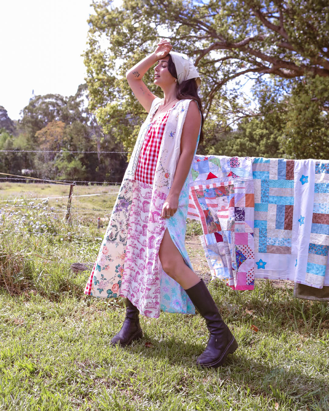 A woman stands outdoors in a grassy field wearing a long, sleeveless patchwork dress with a red and white gingham bodice and a multicolor floral and geometric skirt. A quilted blanket is hanging on a line behind her, with trees in the background.