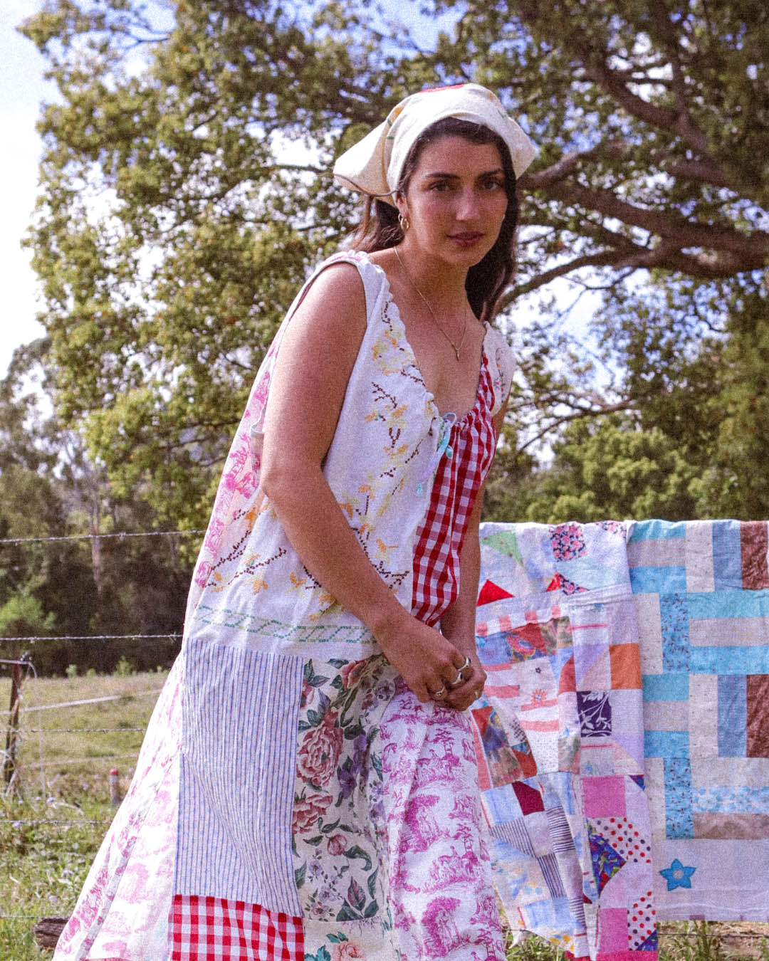 A woman wearing a sleeveless patchwork dress with floral and gingham patterns, standing outdoors next to a colorful patchwork quilt on a line.