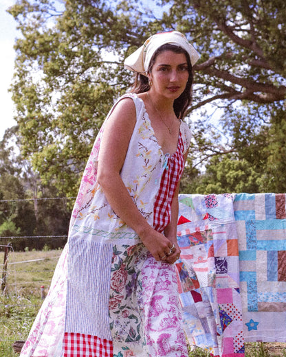 A woman wearing a sleeveless patchwork dress with floral and gingham patterns, standing outdoors next to a colorful patchwork quilt on a line.