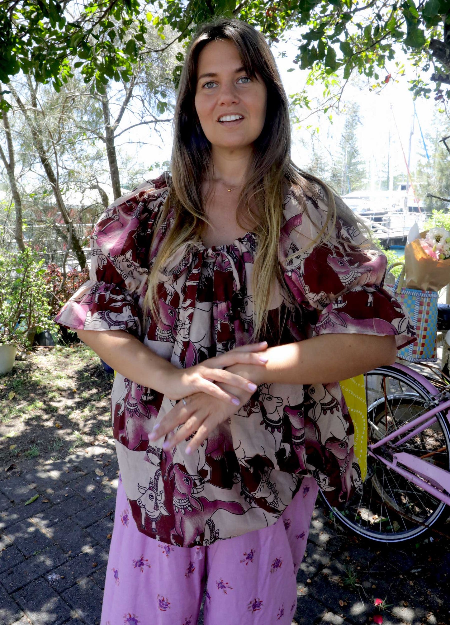 Woman wearing a pink and chartreuse handblock patterned top with holy cow motif and purple pants standing outdoors with trees and plants in the background. A perfect Christmas Fashion Outfit and Black Friday Sale