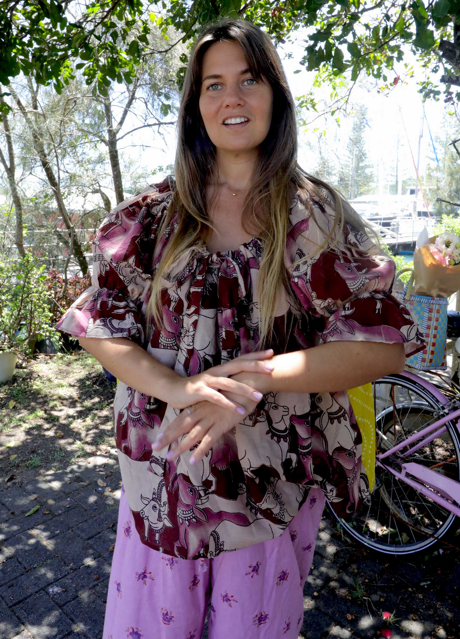 Woman wearing a pink and chartreuse handblock patterned top with holy cow motif and purple pants standing outdoors with trees and plants in the background. A perfect Christmas Fashion Outfit and Black Friday Sale