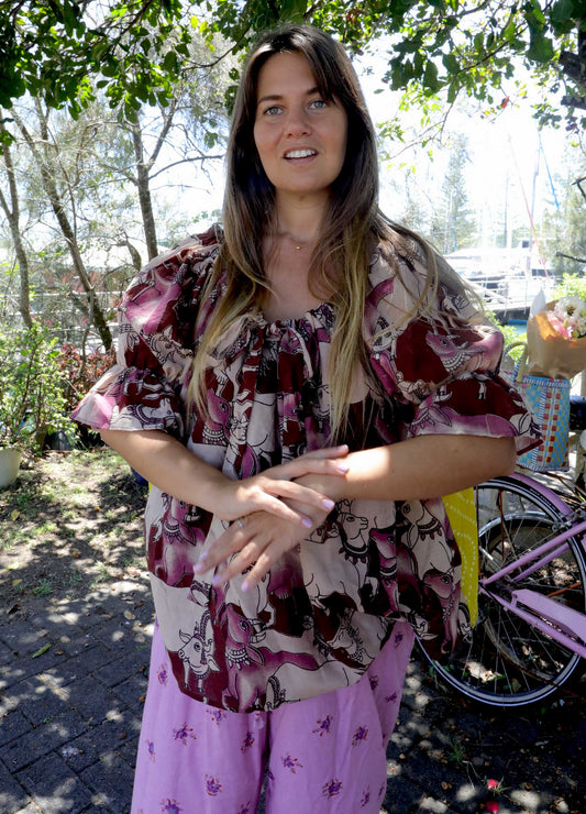 Woman wearing a pink and chartreuse handblock patterned top with holy cow motif and purple pants standing outdoors with trees and plants in the background. A perfect Christmas Fashion Outfit and Black Friday Sale