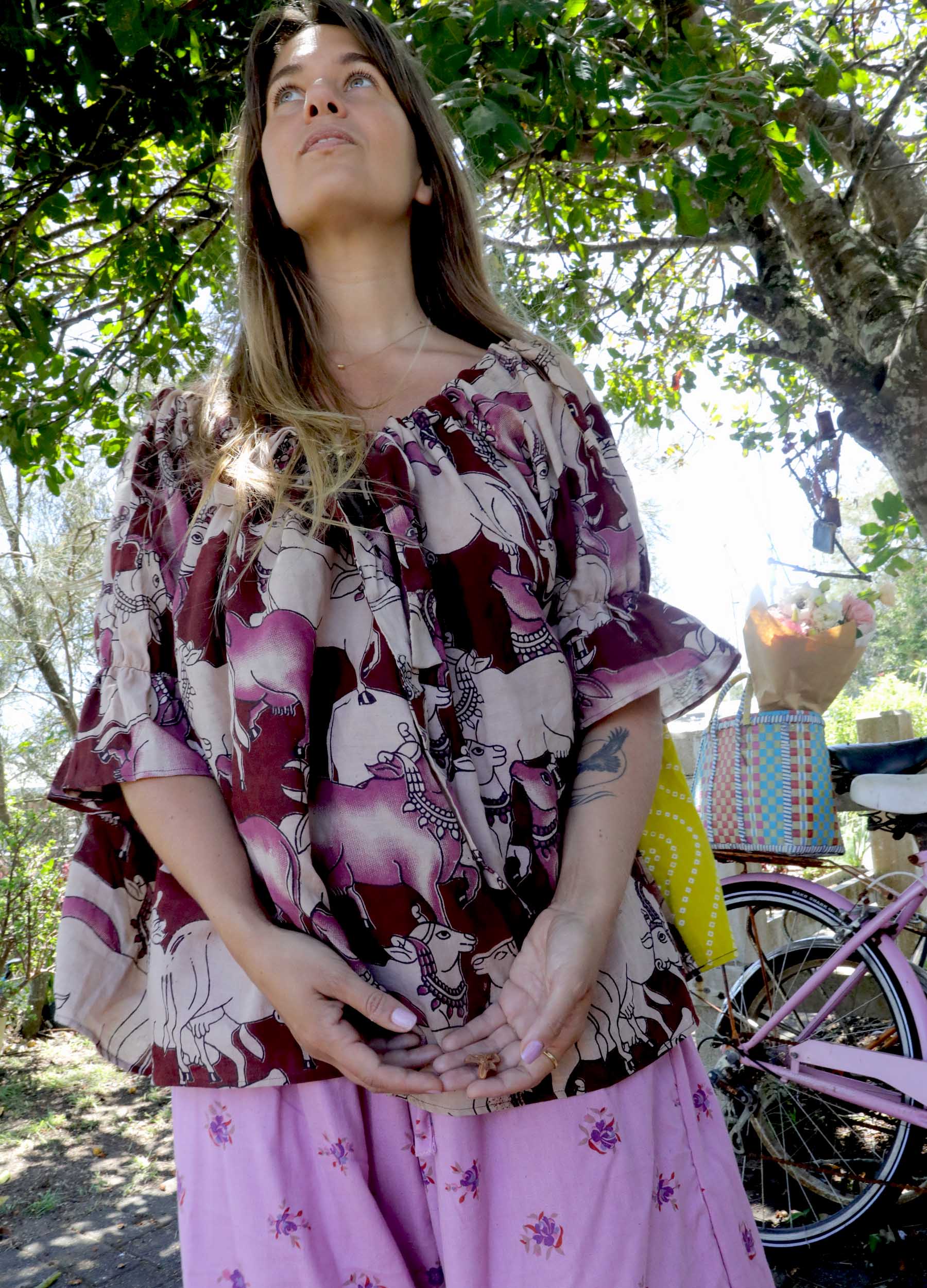 Woman wearing a pink and chartreuse handblock patterned top with holy cow motif and purple pants standing outdoors with trees and plants in the background. A perfect Christmas Fashion Outfit and Black Friday Sale