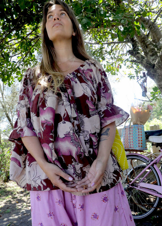 Woman wearing a pink and chartreuse handblock patterned top with holy cow motif and purple pants standing outdoors with trees and plants in the background. A perfect Christmas Fashion Outfit and Black Friday Sale