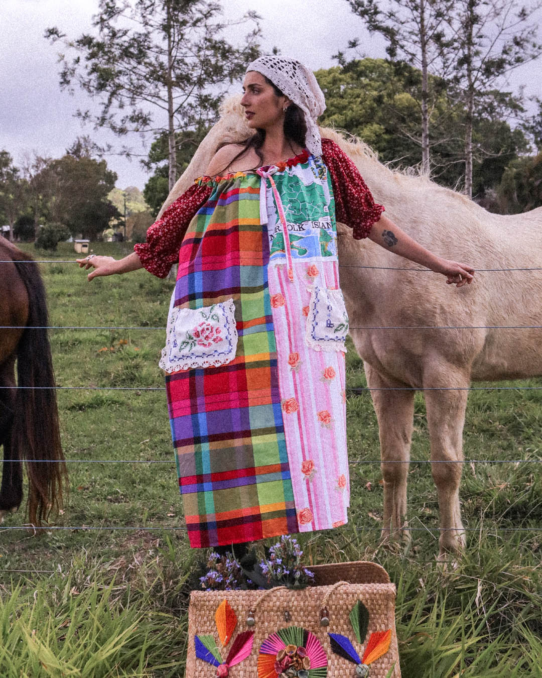 A woman wearing a colorful patchwork maxi dress and a white lace headscarf stands in a grassy field with a light-colored horse behind her. The dress features multiple vibrant plaid and floral patterned panels, red puff sleeves, and decorative white lace pockets.