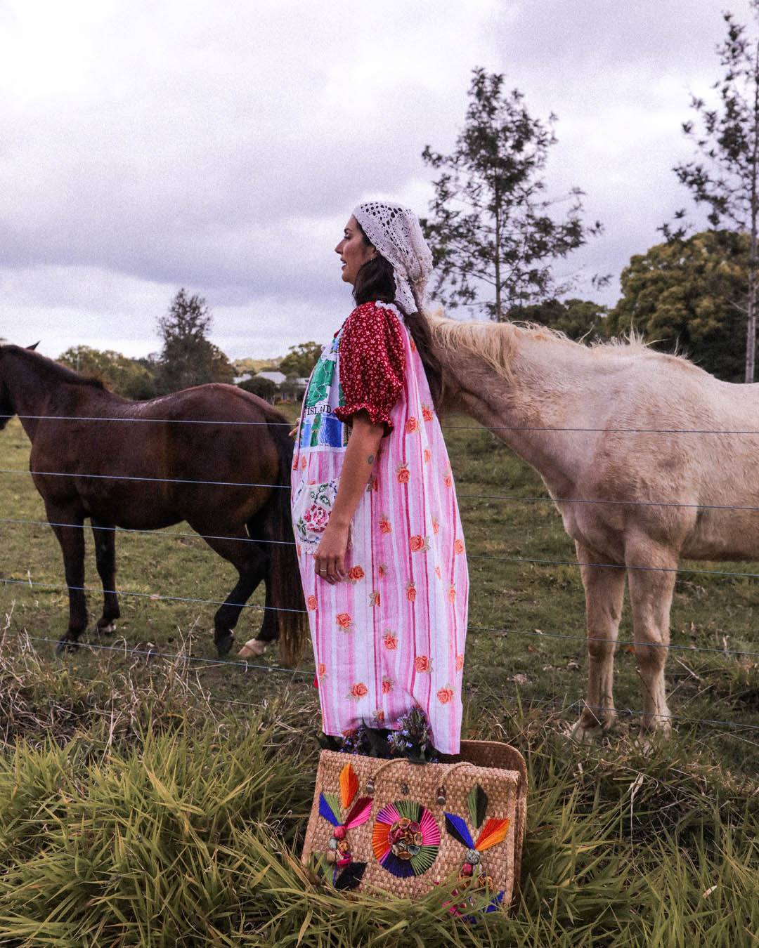 A woman in a one size one of a kind colorful embroidered dress and white lace headscarf stands in a grassy field with two horses behind a wire fence. A woven tote bag with colorful embroidered birds sits on the grass in the foreground.