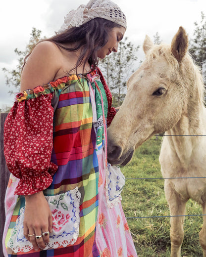 A woman wearing a colorful patchwork bohemian dress and a white crochet headband poses outdoors next to a light-colored horse.