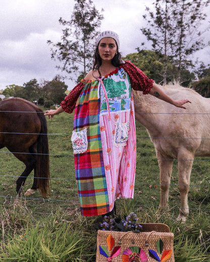 A woman stands outdoors in a grassy field between two horses, wearing a colorful patchwork dress with red puff sleeves and a white headband. A woven tote bag with rainbow feathers sits in the foreground.