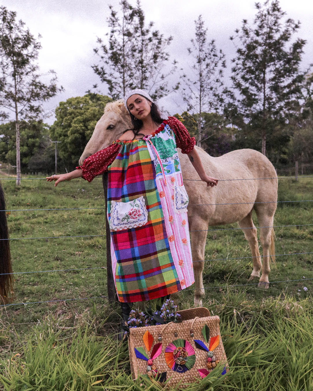 A woman wearing a colorful patchwork dress stands in a grassy field next to a light-colored horse, with a woven tote bag in the foreground.