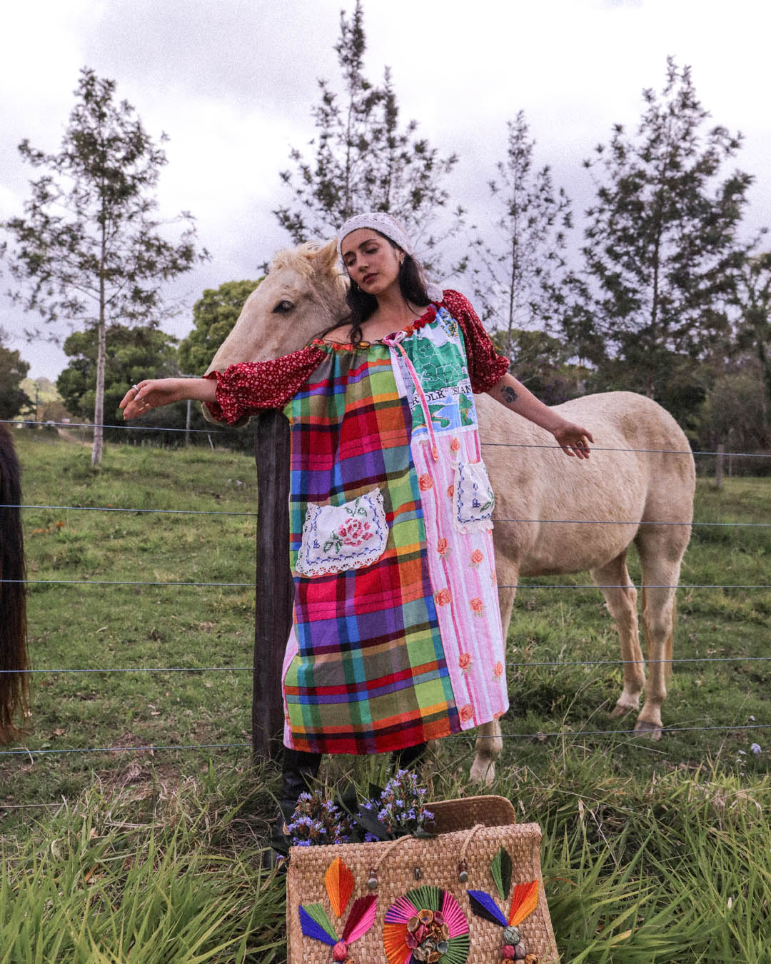 A woman wearing a colorful patchwork apron dress stands outdoors in a grassy field next to a light-colored horse. The dress features a vibrant multicolor plaid and floral pattern with red polka-dot sleeves.