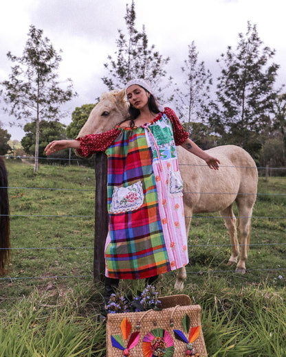 A woman wearing a colorful patchwork apron dress stands outdoors in a grassy field next to a light-colored horse. The dress features a vibrant multicolor plaid and floral pattern with red polka-dot sleeves.