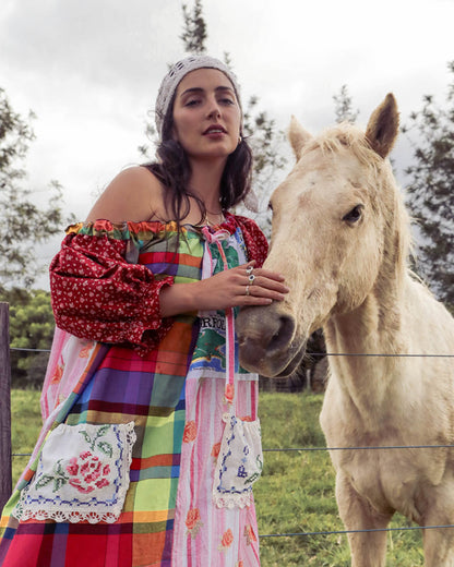 A woman wearing a colorful one of a kind one size patchwork dress and white crochet lace head scarf stands beside a light-colored horse outdoors, with trees and a fence visible in the background.