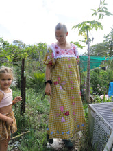 Load image into Gallery viewer, Mother and child standing in a garden with greenery around. Mother is wearing mustard checkered dress with heart applique patterns
