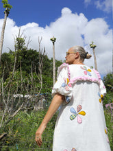 Load image into Gallery viewer, Woman wearing a white dress with colorful floral applique embroidery patterns in a tropical setting.