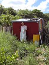 Load image into Gallery viewer, Person in a white dress standing in front of a red shed with greenery around.