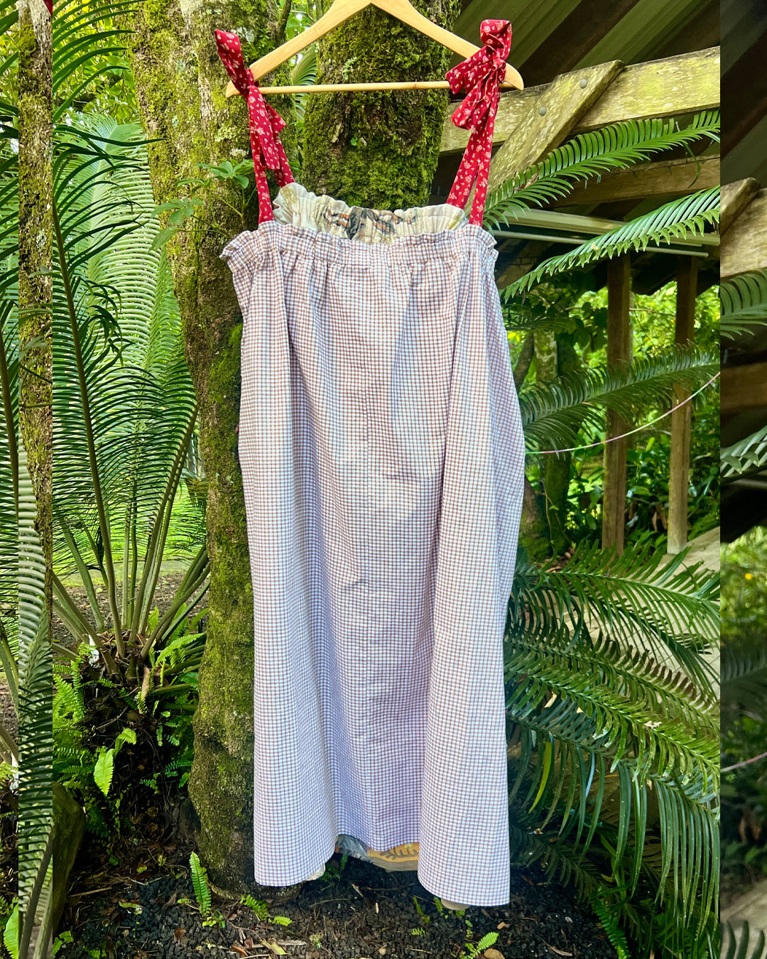 Brown gingham check dress with red floral tie straps hanging on a wooden hanger against a lush green forest background.
